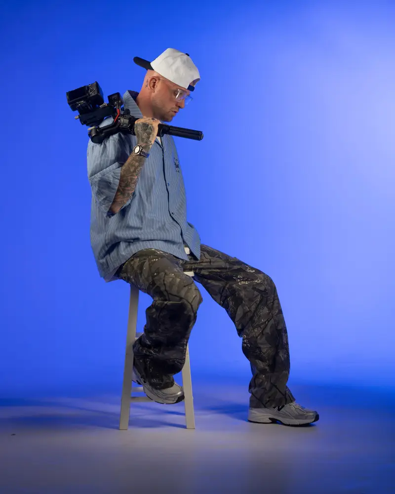 Chris Dodd seated on a studio stool with a camera rig on his shoulder against a blue backdrop